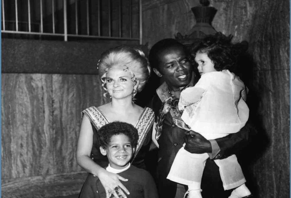 A vintage black-and-white photo of Lou Rawls holding a young child, accompanied by Ceci Rawls and another smiling child, taken at a formal event.