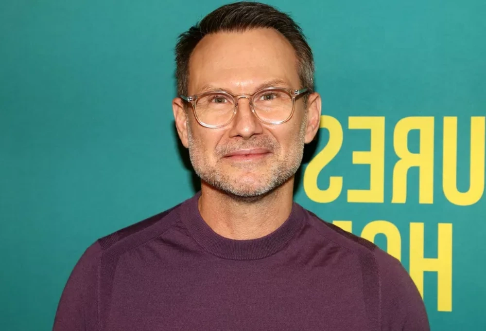 Jaden Christopher Haddon Slater's at a promotional event, wearing glasses and a purple sweater, smiling against a teal and yellow background