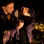 Couple sharing an intimate moment outdoors at night, wrapped in a blanket with string lights in the background