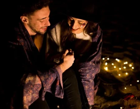 Couple sharing an intimate moment outdoors at night, wrapped in a blanket with string lights in the background