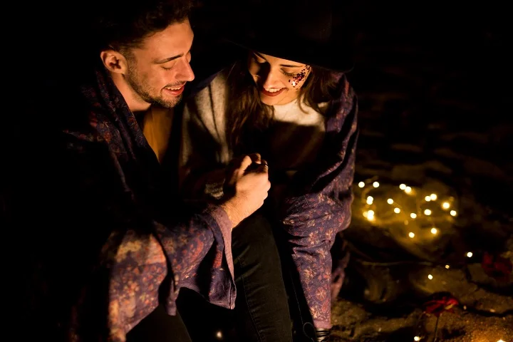 Couple sharing an intimate moment outdoors at night, wrapped in a blanket with string lights in the background