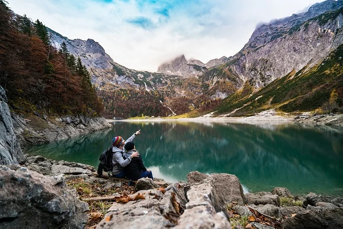 Two people sitting on rocks by a serene mountain lake, surrounded by towering peaks