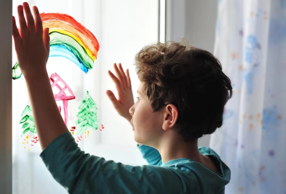 A young boy with curly hair touching a window with a colorful rainbow drawing, symbolizing creativity and self-expression in With Autism.