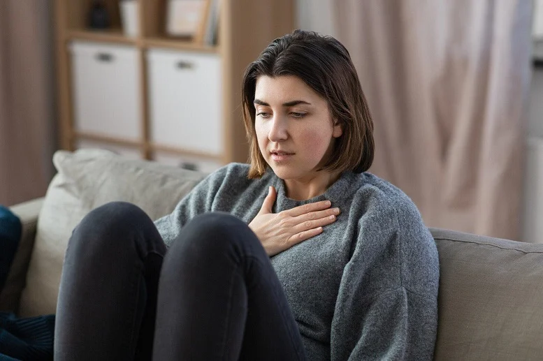 Young woman experiencing shortness of breath after eating, sitting on a couch with her hand on her chest.