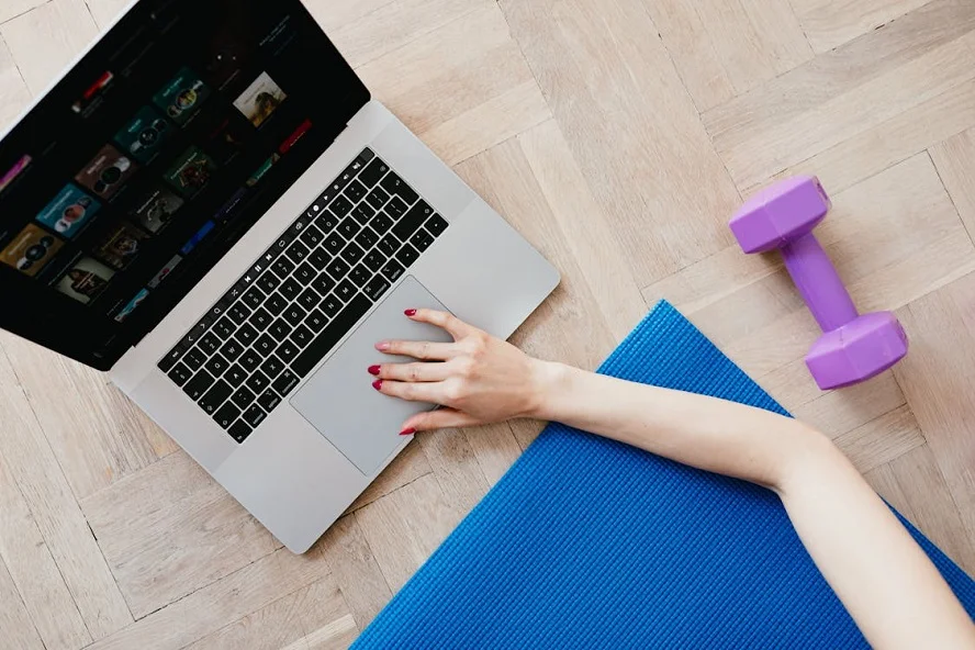 A sports mat, laptop, and dumbbell on a wooden floor, representing personalized fitness tracking and wellness goals.