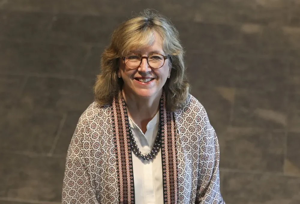 Joseph Renzulli Wife a patterned shawl over a white blouse, accessorized with a beaded necklace, standing indoors on a tiled floor.