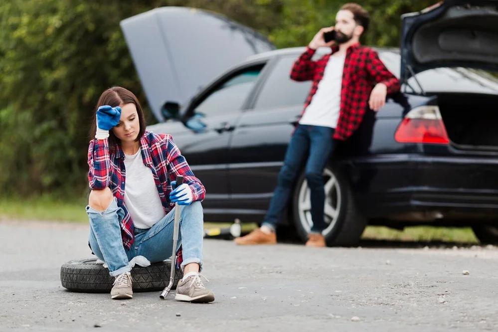 A frustrated woman sitting next to a damaged car while a man calls for help, showing the challenges of life after a car accident.